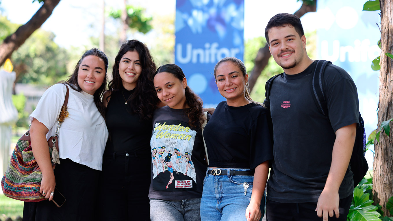 5 alunos: 4 mulheres e 1 homem, em área verde na frente de uma placa escrito 