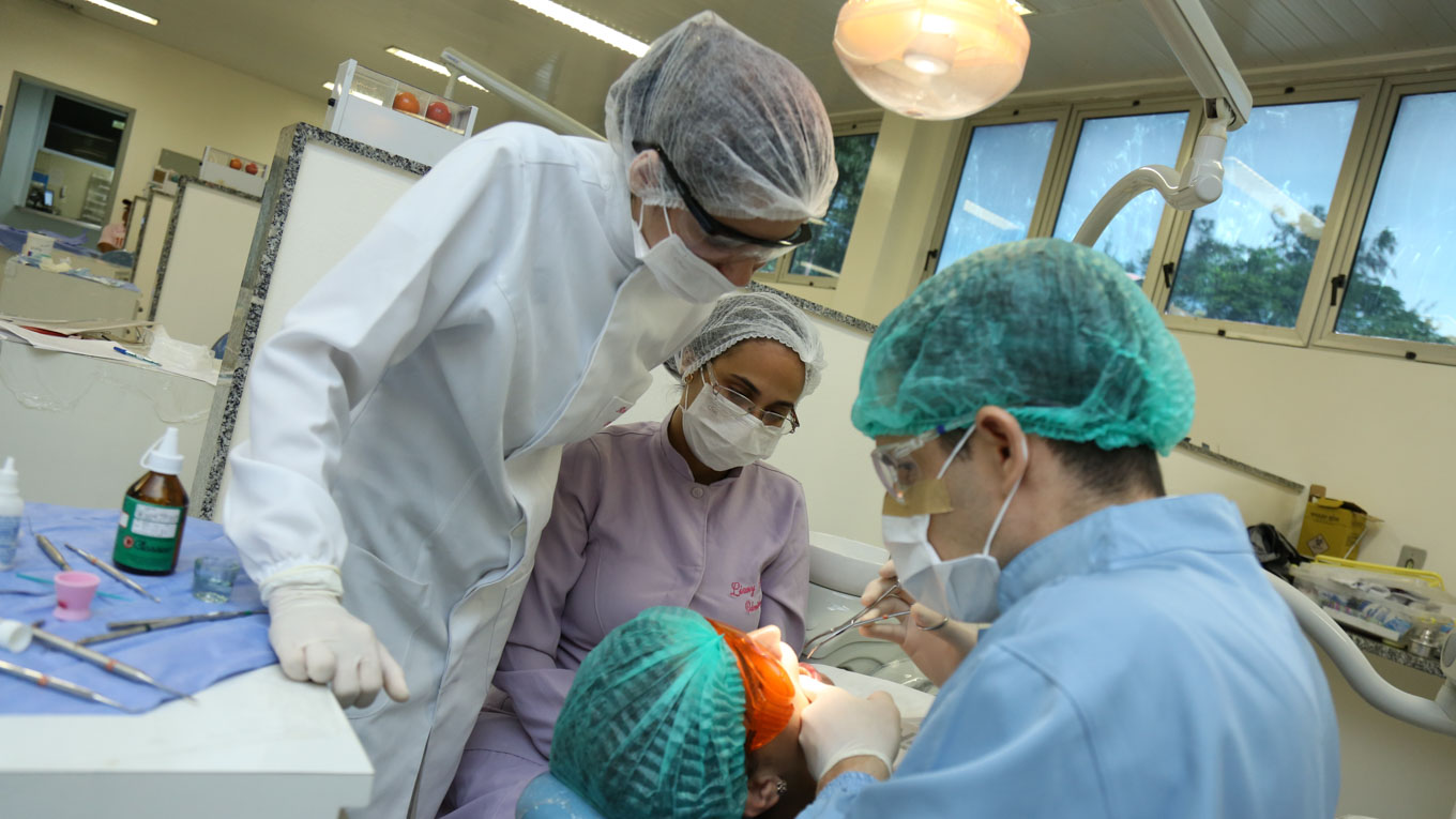 Três profissionais de Odontologia realizam procedimento cirúrgico num paciente.