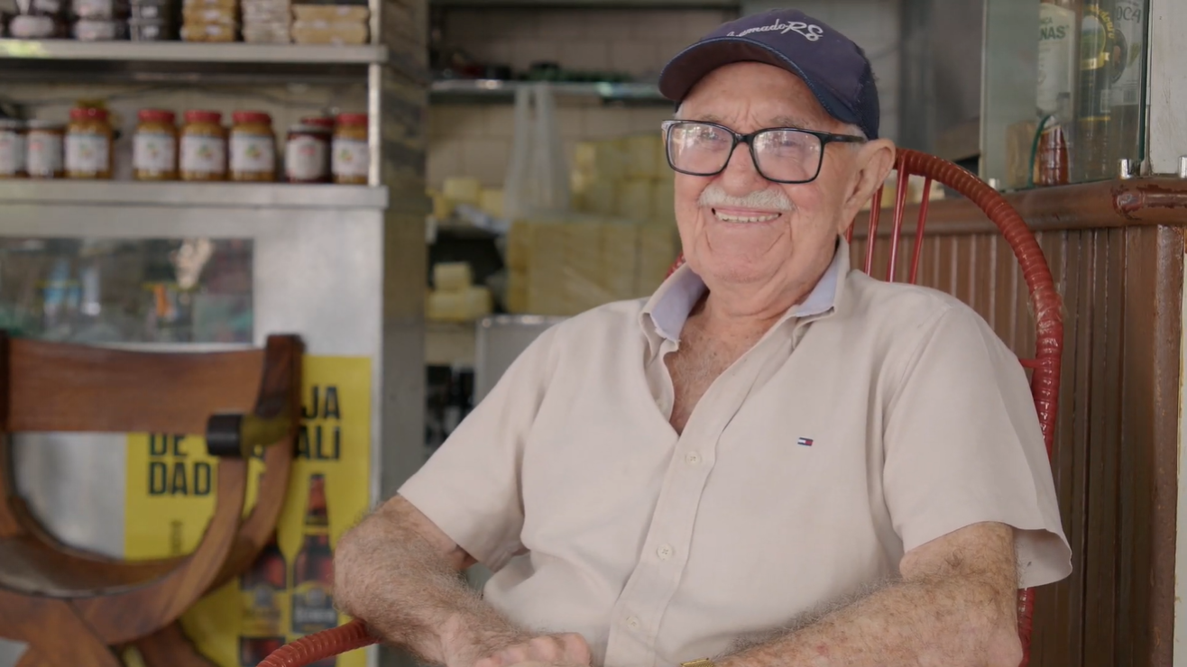 Raimundo do Queijo sorrindo na sua clássica cadeira de balanço vermelha em seu estabelecimento (Foto: Reprodução/TV Unifor)