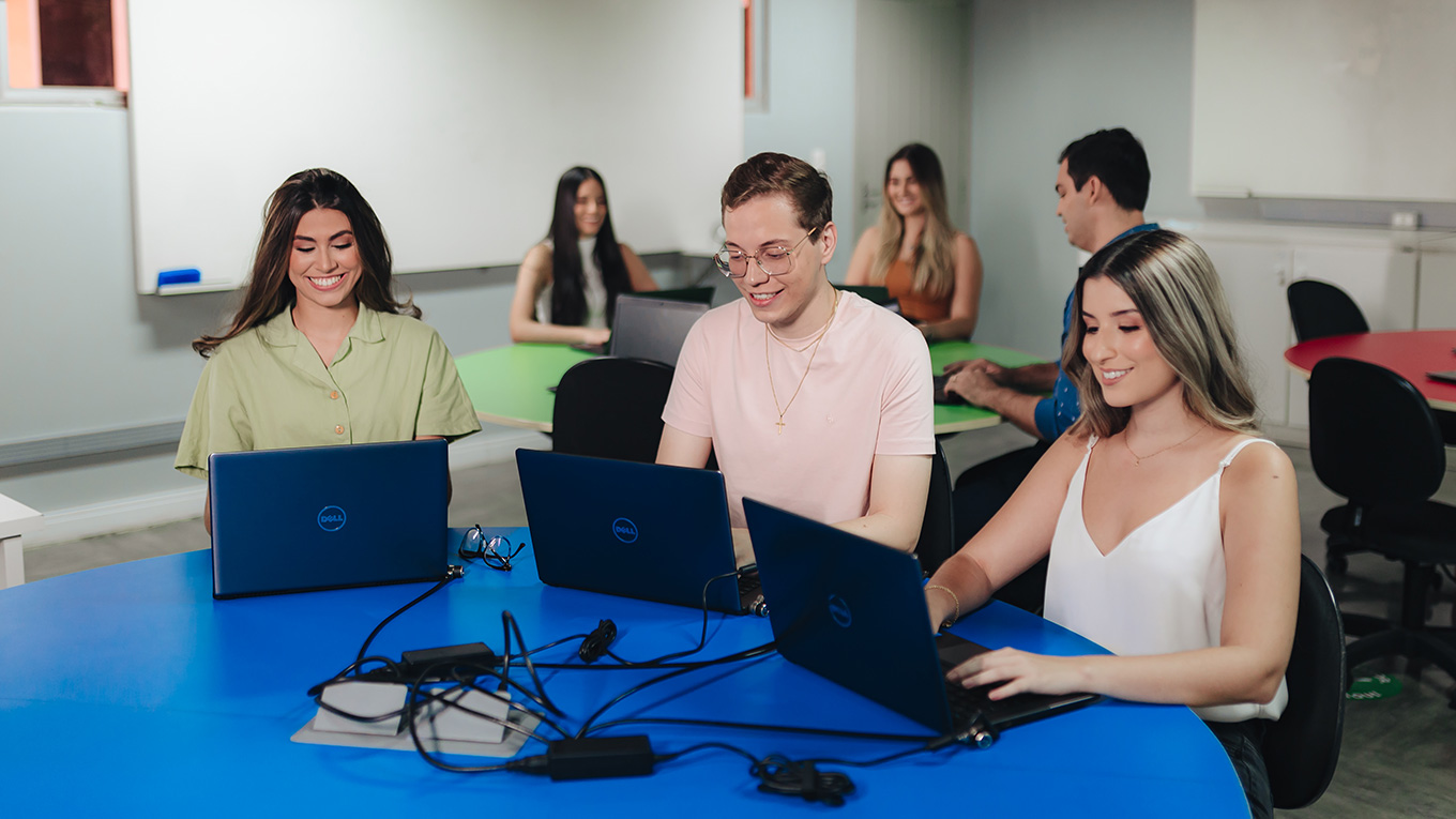 Jovens, sentados ao redor de uma mesa, manipulam seus notebooks