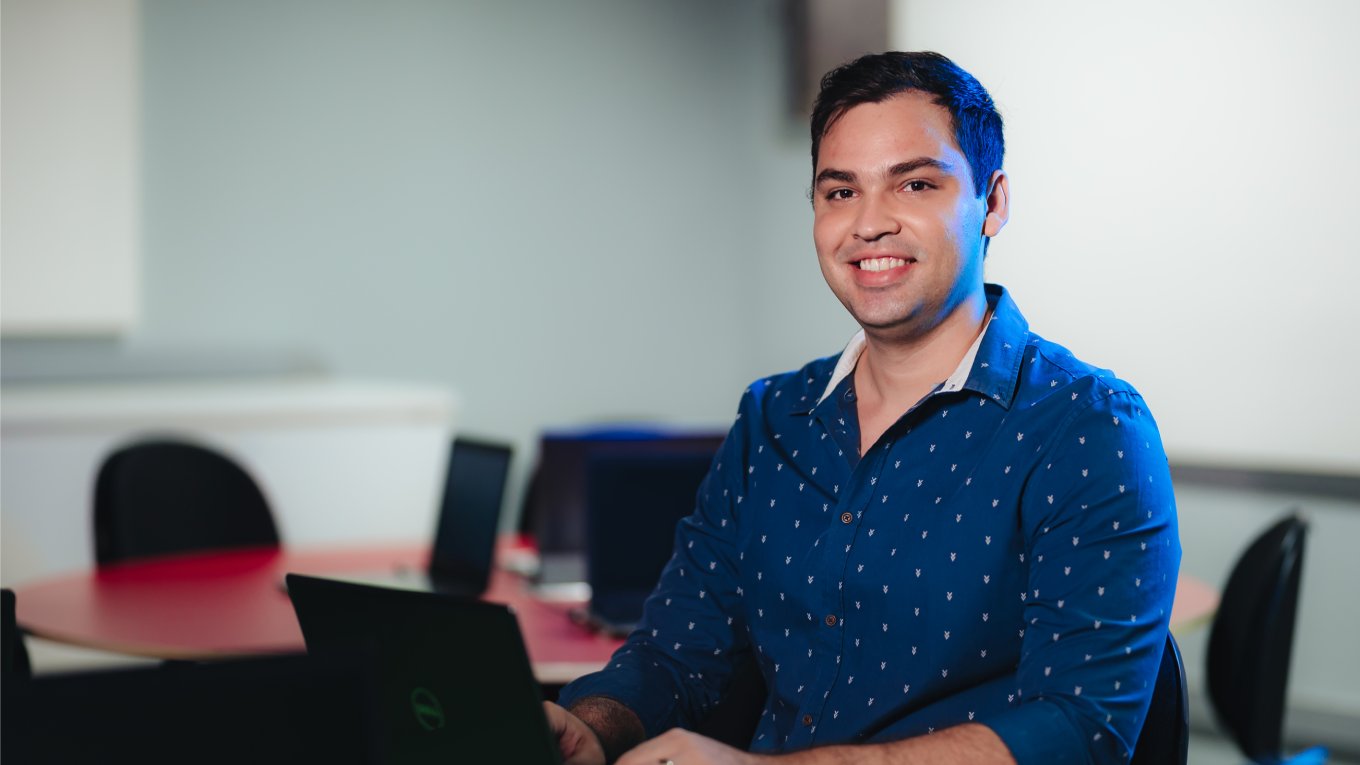 Jovem está sentado com notebook à sua frente; ele tem cabelo curto e usa camisa na cor azul