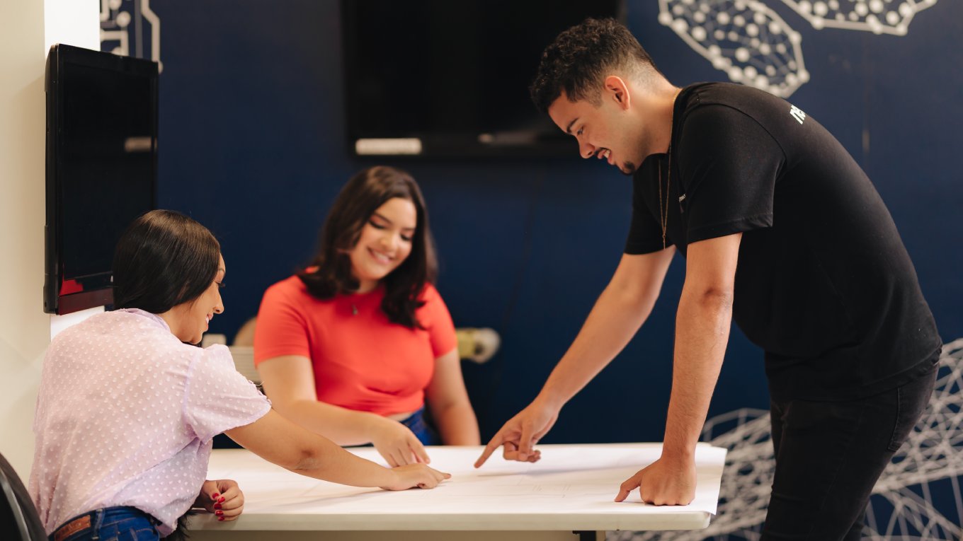 Duas jovens apontam para um documento sobre a mesa; as duas estão sentadas e sorriem